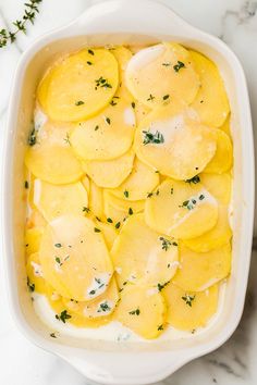 a casserole dish filled with potatoes and cheese on a marble countertop next to herbs