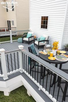 a deck with chairs, table and flowers on the grass next to a white house