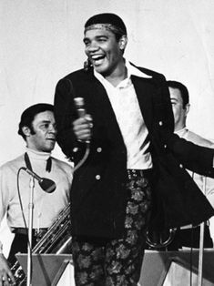 a black and white photo of a man singing into a microphone with other men in the background