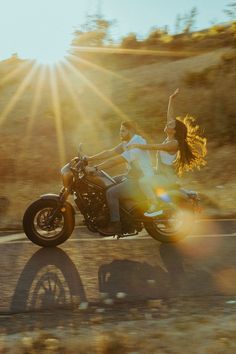 a man and woman riding on the back of a motorcycle down a country road at sunset