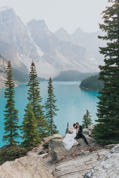 a bride and groom are sitting on the edge of a cliff overlooking a mountain lake