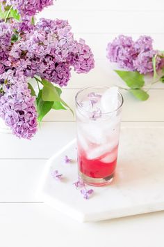 a glass filled with liquid next to purple flowers on a white tray and green leaves