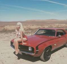 a woman sitting on top of a red car in the middle of desert with her legs crossed