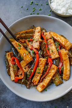 a white bowl filled with tofu and peppers next to rice on a table top