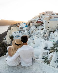 two people sitting on the edge of a cliff looking at the ocean and white buildings