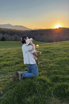 a woman holding a baby in her arms while sitting on the grass with mountains in the background