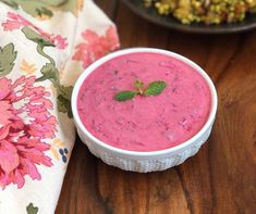 a white bowl filled with pink liquid on top of a wooden table next to a plate of food