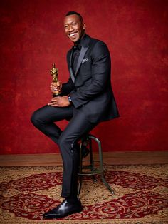 a man in a tuxedo sitting on a stool holding an oscar statue and smiling