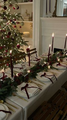 a table set for christmas dinner with lit candles and plates on it, next to a decorated christmas tree