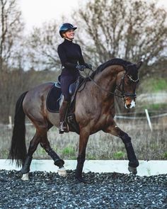 a woman riding on the back of a brown horse across a gravel covered field with trees in the background