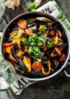 a pan filled with mussels and vegetables on top of a table next to wine glasses