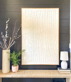 two vases with plants sit on a shelf in front of a framed art piece