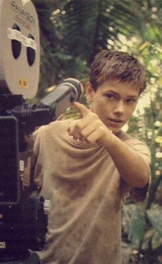 a young boy pointing at the camera in front of some plants and trees with his thumb up