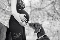 a black and white photo of a pregnant woman with her dog in front of trees