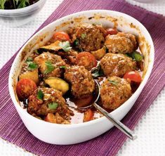 meatballs and vegetables in a white dish with spoons on a purple place mat