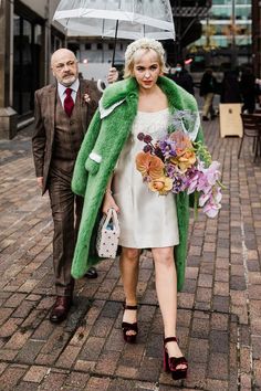 a woman in a white dress and green coat holding an umbrella while walking down the street