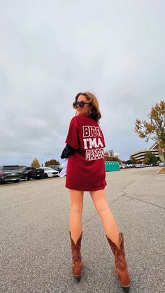 a woman in a red shirt dress and cowboy boots is standing on the street with her legs crossed