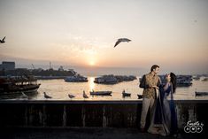 a man and woman standing next to each other near the water with birds flying in the sky