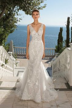 a woman in a white wedding dress standing on steps near the ocean with trees and bushes