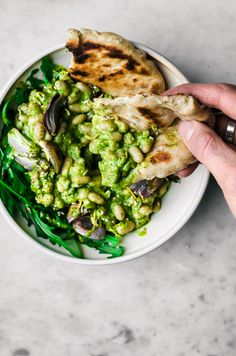 a person holding a piece of food over a bowl filled with green vegetables and pita bread