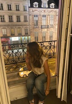 a woman sitting on a window sill looking out at the street from her apartment