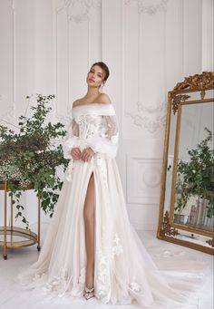 a woman wearing a white wedding dress with long sleeves and flowers on the skirt, standing in front of a mirror