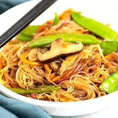 a white bowl filled with noodles and vegetables next to chopsticks on a blue cloth