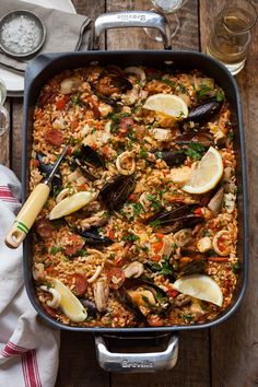 a pan filled with rice and mussels on top of a wooden table next to wine glasses