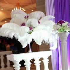 a tall white vase filled with flowers and feathers on top of a table next to a purple curtain