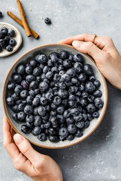 two hands holding a bowl of blueberries with cinnamon sticks on the side next to it