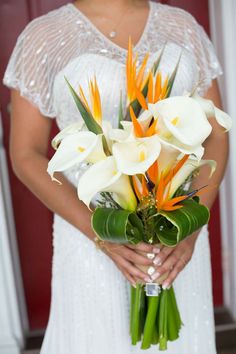 a woman in a white dress holding a bouquet of orange and white flowers with greenery