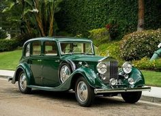 an old green car is parked on the side of the road in front of some bushes