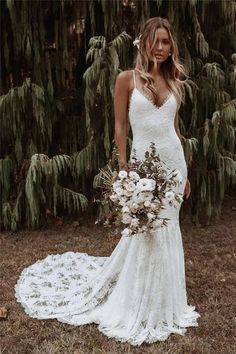 a woman in a white wedding dress standing next to some trees and holding a bouquet