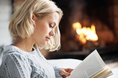 a woman reading a book in front of a fire place with her head turned to the side