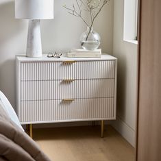 a white dresser sitting next to a bed with a lamp on top of it and a vase filled with flowers