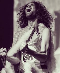 a man with long curly hair and beards singing into a microphone while wearing a furry vest