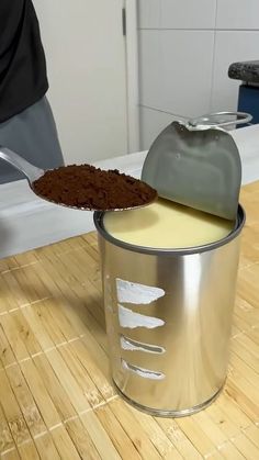 a person holding a spoon over a paint can with some brown stuff in it on top of a wooden floor