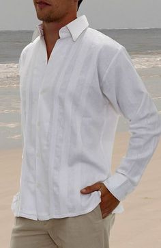 a man standing on the beach with his hands in his pockets, wearing a white shirt and khaki pants