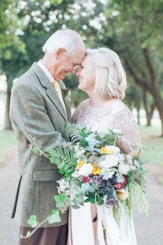 an older man and woman standing next to each other in front of some trees with flowers