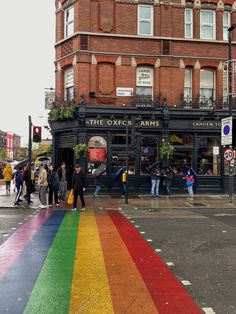 people are standing on the sidewalk in front of an old building with a rainbow painted crosswalk