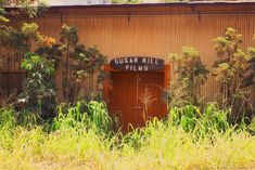 a red door in front of a building with tall grass and trees around it that reads sugar mill films