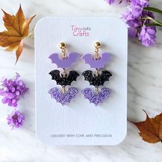 the bat earrings are purple and black with glitter accents on them, sitting next to some flowers