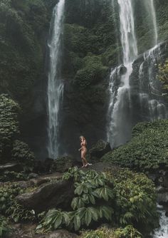 a naked woman standing in front of a waterfall with lush green foliage and trees around her