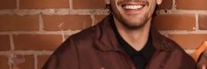 a man is smiling and holding an orange object in front of his face while standing next to a brick wall