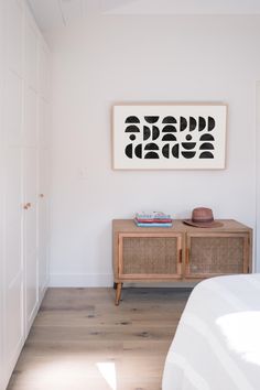 a white room with a wooden sideboard and artwork on the wall, along with a bed