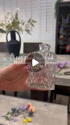 a person holding up a glass bottle with flowers on the table in front of them