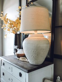 a white vase sitting on top of a dresser next to a mirror and lamp shade