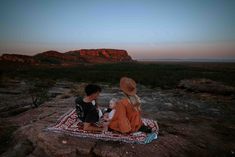 two children sitting on a blanket looking at the sunset in the desert with mountains behind them