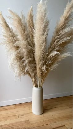 a white vase filled with dry grass on top of a wooden floor