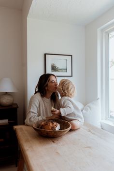 a woman and child sitting at a table in front of a window with a bowl of food on it